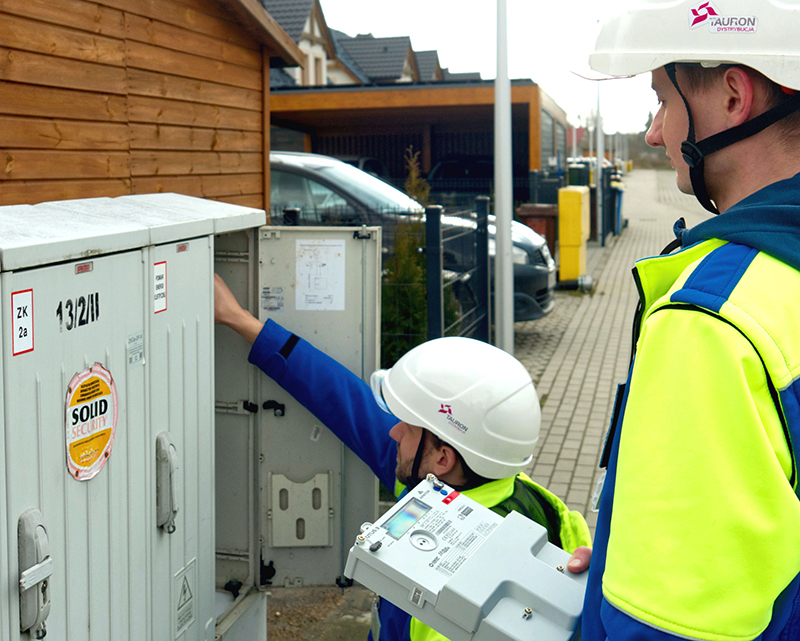 dwóch elektromonterów wymienia licznik energii elektrycznej na nowy w skrzynce znajdującej się obok domu